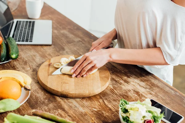 Vista cortada da jovem mulher cortando bananas com faca à mesa na cozinha — Fotografia de Stock