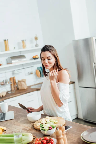 Fröhliche junge Frau hält Messer in der Hand und steht neben Tisch in Küche — Stockfoto