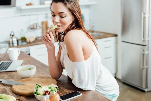 Charming girl sitting at table and eating salad in kitchen — Stock Photo