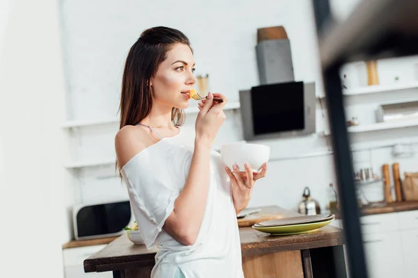 Dreamy woman holding bowl and eating cut fruit in kitchen — Stock Photo