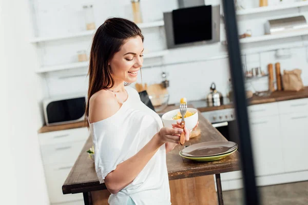 Souriant jeune femme tenant bol et manger des fruits coupés dans la cuisine — Photo de stock