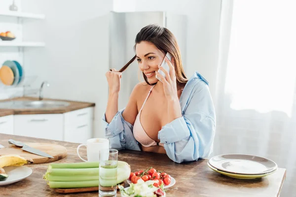 Sexy girl in bra and short sitting at table and talking on smartphone — Stock Photo