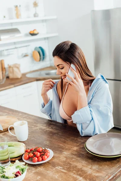 Sexy girl in bra and short sitting at table and talking on smartphone — Stock Photo