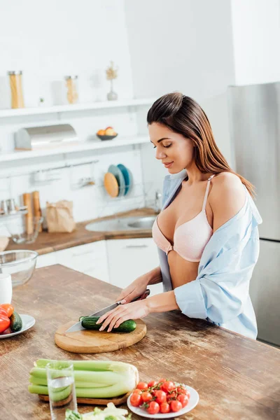 Sexy woman in bra and shirt cutting cucumber with knife in kitchen — Stock Photo