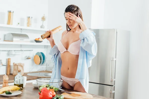 Tired woman in lingerie and shirt holding spice grinder while cooking vegetable salad — Stock Photo