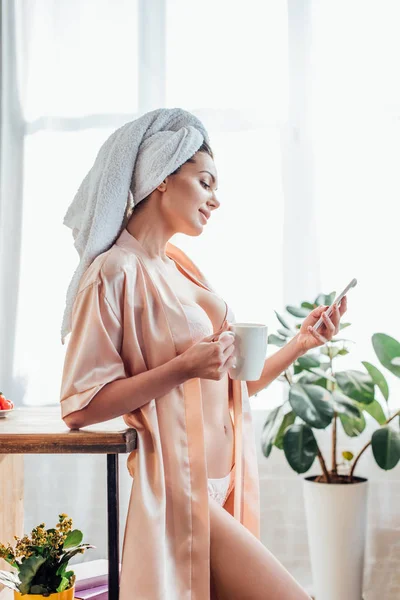 Sexy chica en lencería y casa con toalla en la cabeza sosteniendo taza de café y el uso de teléfono inteligente en la cocina - foto de stock