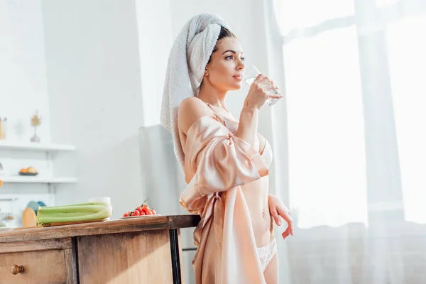 Menina sexy em lingerie branca e housecoat segurando vidro de água na cozinha — Fotografia de Stock