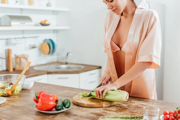 Cropped view of sexy woman in housecoat with towel on head cutting celery with knife in kitchen — Stock Photo