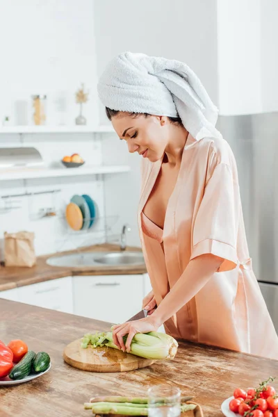 Mulher sexy em housecoat com toalha na cabeça cortando aipo com faca na cozinha — Fotografia de Stock