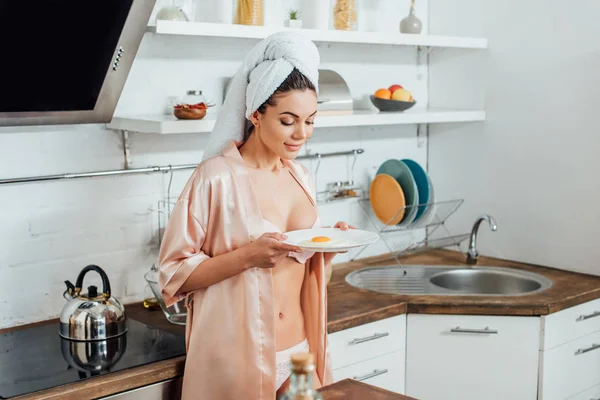 Сексуальная женщина в халате держит тарелку с жареным яйцом на кухне — стоковое фото