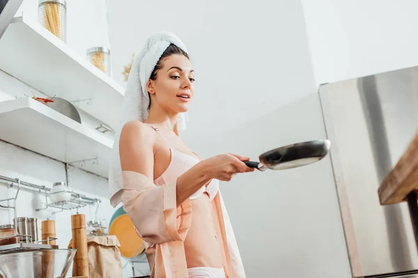 Vue à angle bas de fille sexy en housecoat avec serviette sur la tête tenant poêle à frire dans la cuisine — Photo de stock