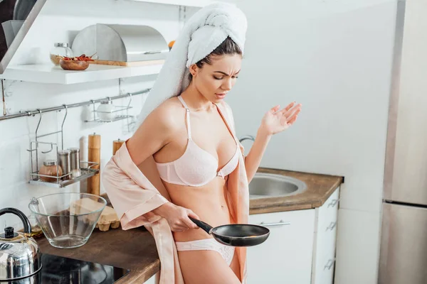 Dissatisfied sexy girl in housecoat with towel on head holding frying pan in kitchen — Stock Photo