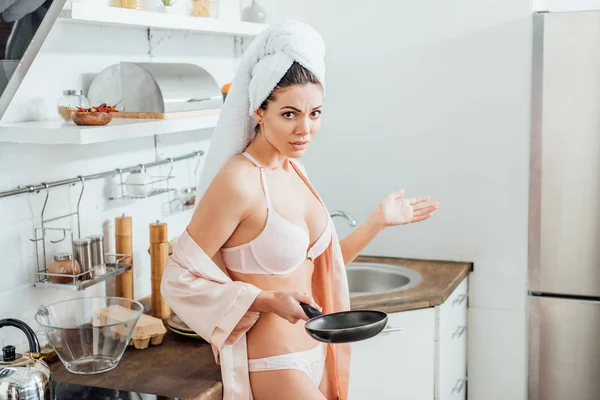 Недовольная сексуальная девушка в халате с полотенцем на голове, держащая сковородку на кухне — стоковое фото