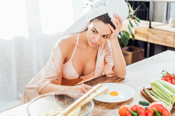 Ragazza pensierosa in casalinga con asciugamano sulla testa seduta a tavola con uovo fritto in cucina — Foto stock