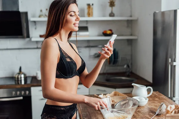 Femme en lingerie noire fouettant des œufs avec fouet et tenant smartphone dans la cuisine — Photo de stock