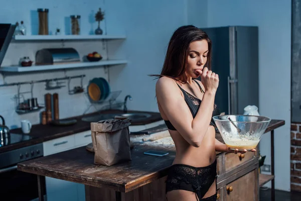 Sexy jovem mulher em lingerie preta lambendo o dedo enquanto chicotadas ovos com batedor na cozinha — Fotografia de Stock