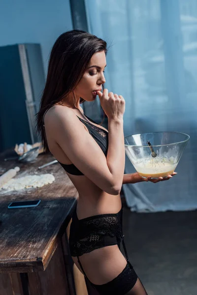Side view of sexy young woman in black lingerie licking finger while whipping eggs with whisk in kitchen — Stock Photo