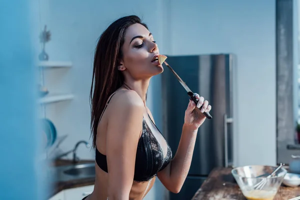 Sexy chica en lencería sosteniendo cuchillo y comiendo manzana en la cocina - foto de stock