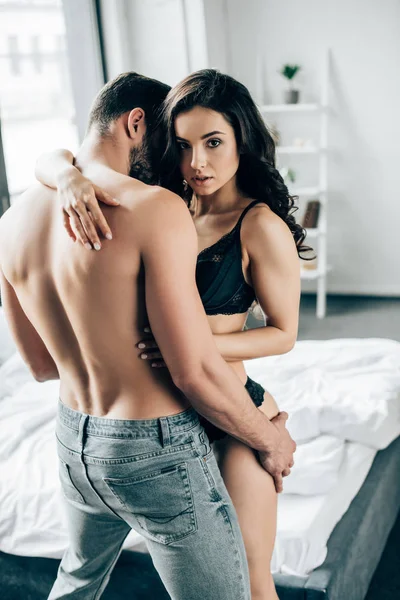 Приваблива брюнетка жінка обіймає без сорочки чоловіка в спальні — стокове фото