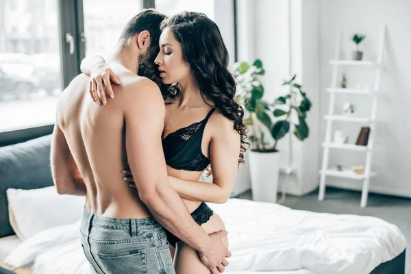 Atractiva mujer morena con los ojos cerrados abrazo de hombre sin camisa en el dormitorio - foto de stock