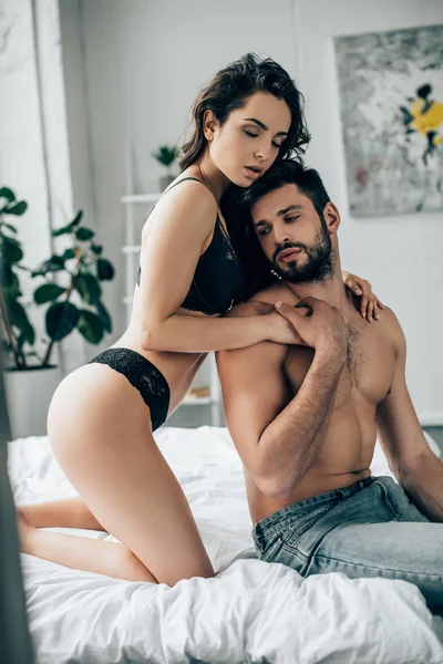 Брюнетка сексуальная женщина в нижнем белье обнимает мужчину без рубашки на кровати — стоковое фото