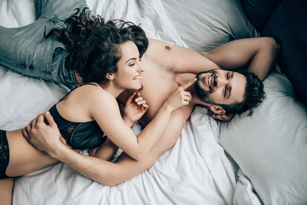 Overhead view of happy bearded man lying with smiling brunette woman in black underwear on bed — Stock Photo