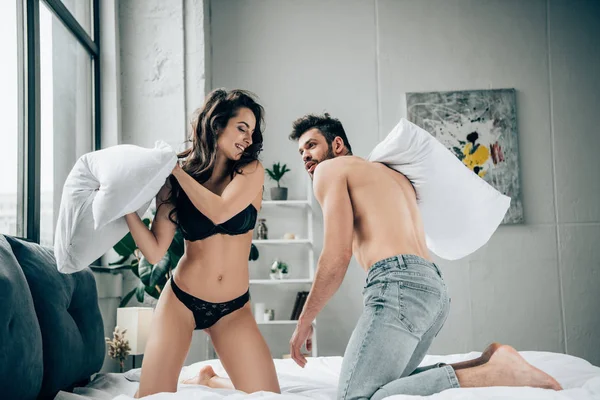 Cheerful brunette woman in lace underwear having pillow fight with muscular boyfriend on bed — Stock Photo