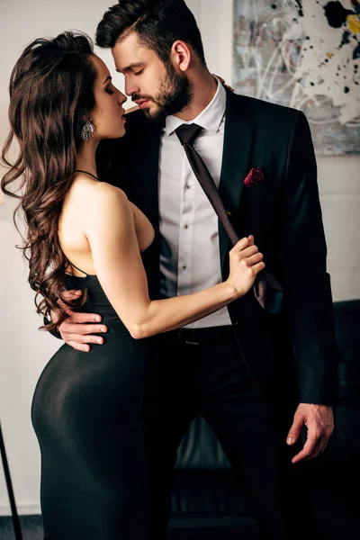 Attractive woman in black dress holding tie of passionate man standing in suit — Stock Photo