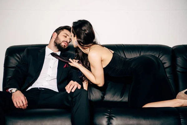 Attractive girl in black dress holding tie of passionate man in suit sitting on sofa — Stock Photo