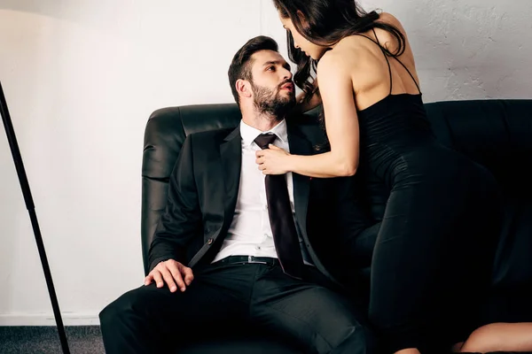 Brunette girl in black dress holding tie of passionate man in suit sitting on sofa — Stock Photo