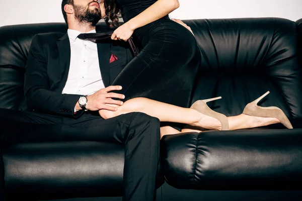 Vue recadrée de fille sexy en robe noire tenant cravate de l'homme barbu assis sur le canapé — Photo de stock