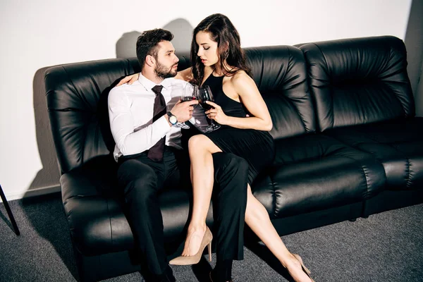 Barbudo hombre y mujer en vestido negro sentado en el sofá y sosteniendo vasos de vino - foto de stock