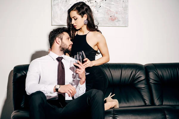 Hombre guapo mirando a la chica en vestido negro mientras está sentado en el sofá y sosteniendo un vaso de vino — Stock Photo