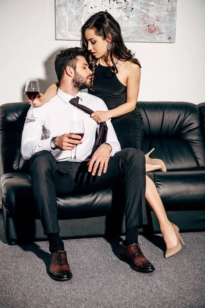 Schöner Mann, der eine Frau im schwarzen Kleid ansieht, während er auf dem Sofa sitzt und ein Glas Wein in der Hand hält — Stockfoto
