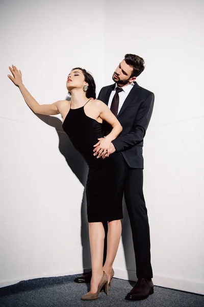 Brünette Frau im schwarzen Kleid hält Händchen mit Mann nahe weißer Mauer — Stockfoto