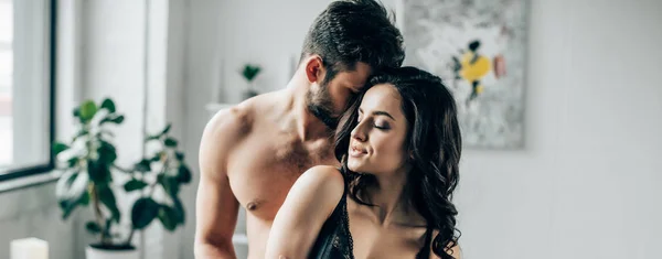 Tiro panorámico del hombre abrazando novia sexy con los ojos cerrados en el dormitorio - foto de stock