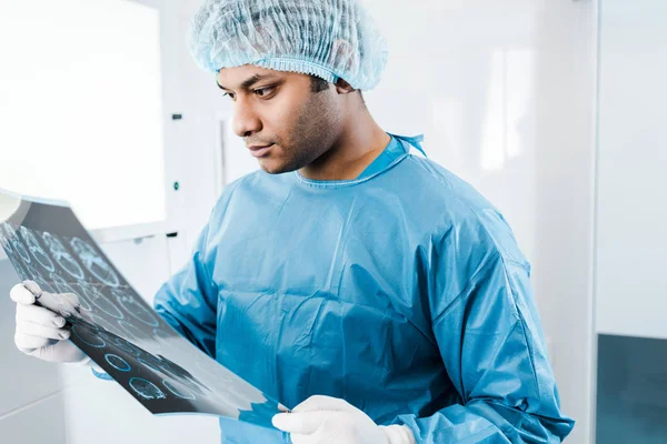 Médico bonito em boné médico e luvas de látex segurando e olhando para raio-x — Fotografia de Stock