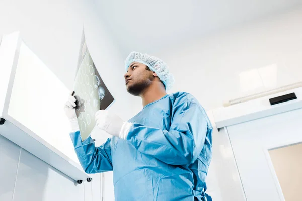 Vista de ángulo bajo del médico en gorra médica y guantes de látex mirando rayos X - foto de stock