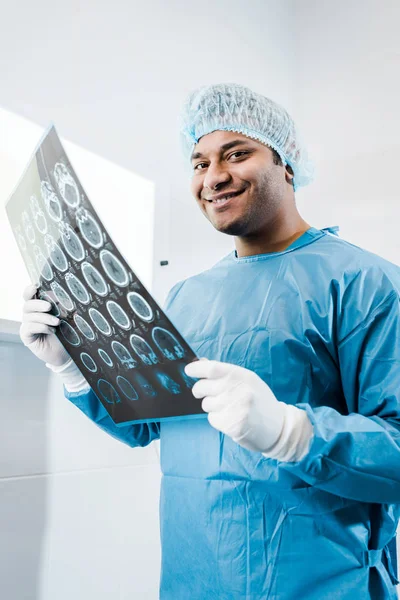 Médico sorridente em uniforme e tampa médica segurando raio-x e olhando para a câmera — Fotografia de Stock
