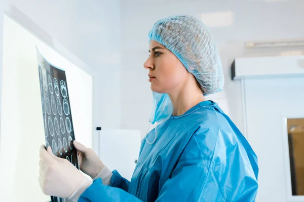 Vue latérale du médecin en casquette médicale et uniforme tenant des rayons X dans la clinique — Photo de stock