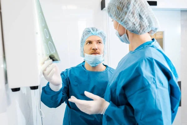 Médicos con uniformes y máscaras médicas hablando de rayos X en la clínica - foto de stock