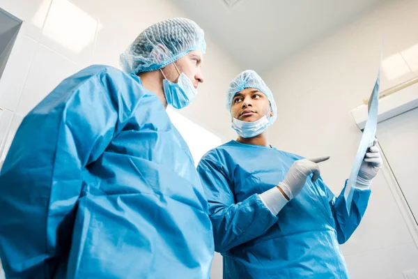 Tiefansicht von gut aussehenden Ärzten in Uniformen und medizinischen Masken, die über Röntgenaufnahmen sprechen — Stockfoto