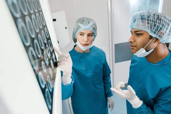 Médicos guapos y hermosos en uniformes y máscaras médicas que señalan con los dedos a la radiografía - foto de stock