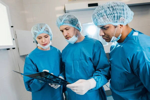 Gut aussehende und schöne Ärzte in Uniformen und medizinischen Masken, die über Röntgenaufnahmen sprechen — Stockfoto