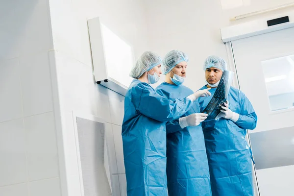Gut aussehende und attraktive Ärzte in Uniformen und medizinischen Masken, die mit dem Finger auf Röntgenbilder zeigen und reden — Stockfoto
