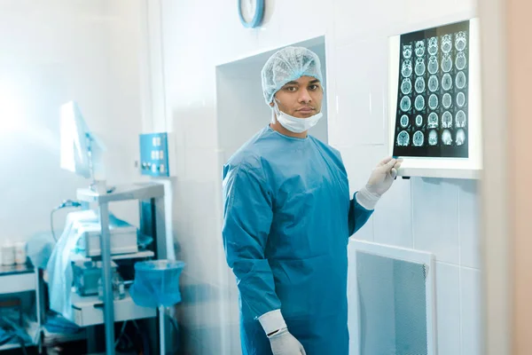 Médico bonito em uniforme e máscara médica segurando raio-x e olhando para a câmera na clínica — Fotografia de Stock