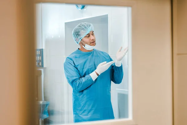 Médico bonito em uniforme e máscara médica colocando em luva de látex e olhando para longe — Fotografia de Stock