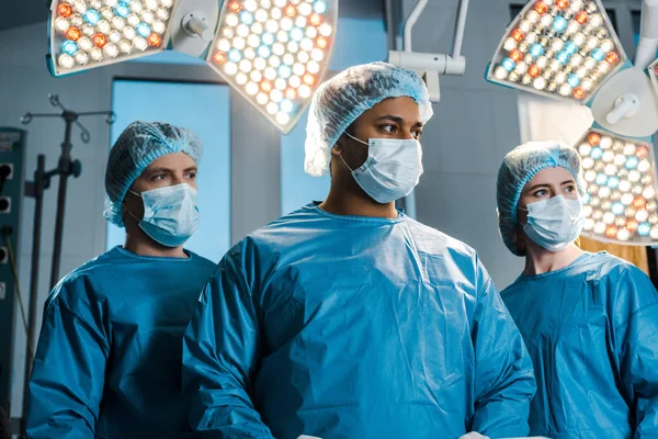 Médicos y enfermeras en uniformes y máscaras médicas mirando hacia otro lado en el quirófano - foto de stock