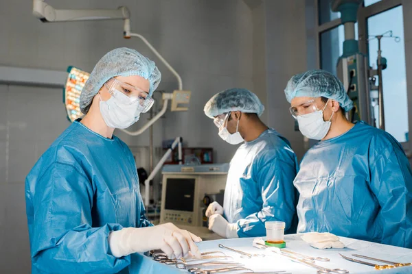 Врачи делают операцию и медсестра в униформе и медицинской шапке давая оборудование — стоковое фото