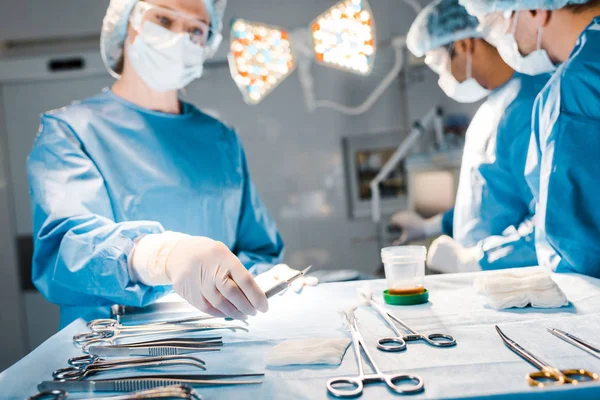 Selective focus of doctors doing operation and nurse in uniform and medical cap holding scalpel — Stock Photo